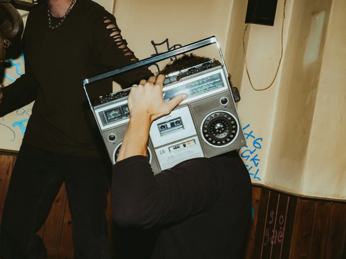 A person in a dimly lit bar carries a silver boombox on one shoulder