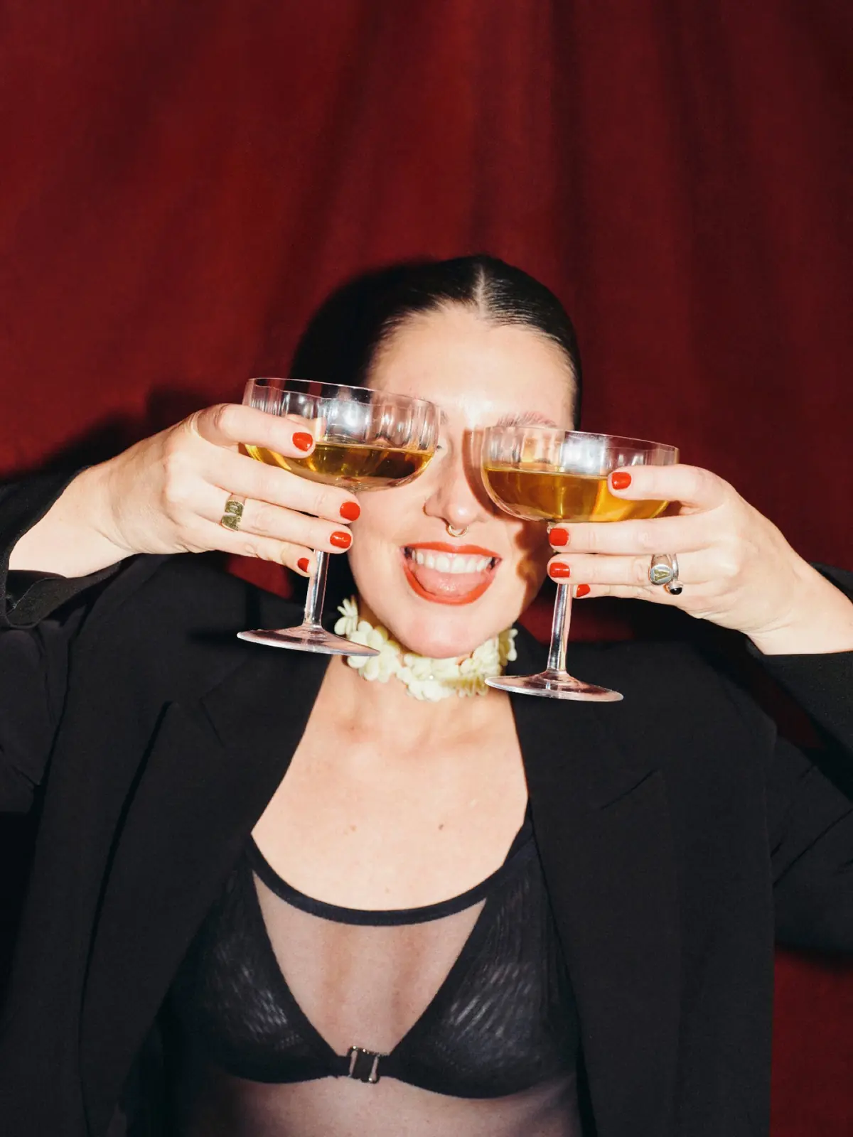 A smiling woman holds two coupe glasses of champagne over her eyes against a red curtain