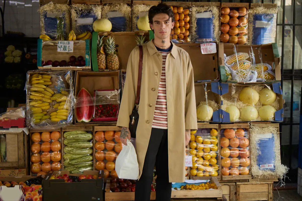 A young man in a camel coat stands in front of a market stall stacked with fruit